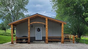 A rustic wooden building with a grey exterior, a door, and a bench outside. - The Lodge at 5J Hunts (Carrollton)