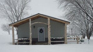 A wooden cabin with a door and a wreath, surrounded by snow-covered trees and a wooden fence. - The Lodge at 5J Hunts (Carrollton)