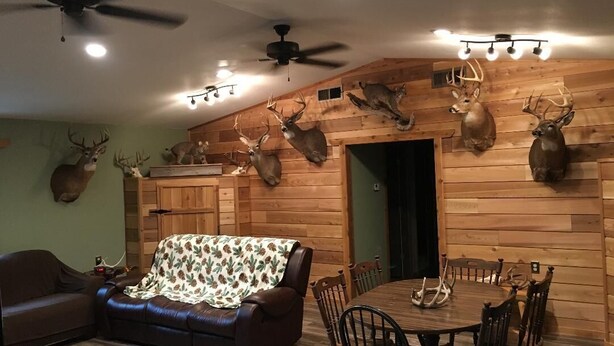 A cozy living room with a wooden wall adorned with deer antlers, a brown leather sofa, a dining table with chairs, and ceiling fans. - The Lodge at 5J Hunts (Carrollton)