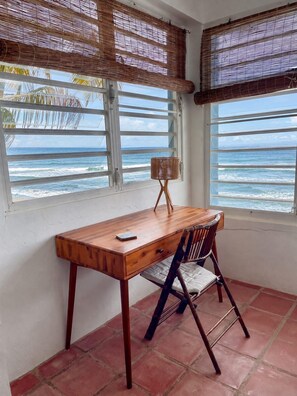A room with a wooden desk and chair, a window with a view of the ocean, and a woven bamboo blind. - BEACH HOUSE W/ POOL (vieques)
