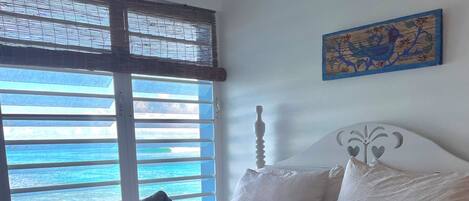 A neatly made bed with a grey bedspread and pillows, a window with blinds, and a wall-mounted artwork.