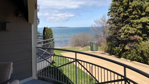 A balcony with a view of the sea, a green lawn, and a tall evergreen tree.
