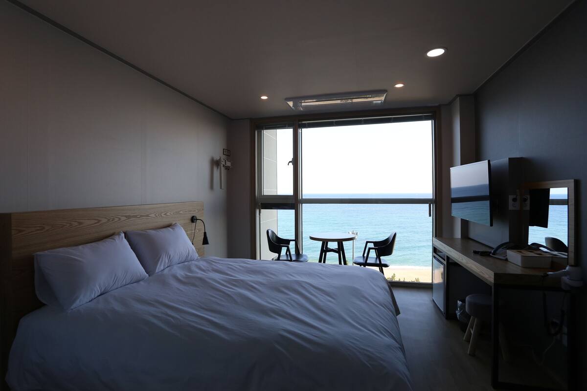 A modern hotel room with a large bed, a desk with a TV, and a view of the sea.