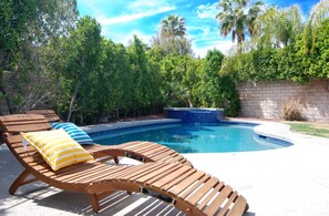 Relax by the serene pool surrounded by lush greenery and palm trees, with a low wall and lounge chairs for a tranquil retreat.