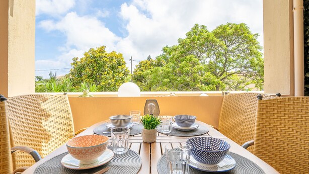 Un espacio comedor al aire libre con una mesa dispuesta para dos, rodeado de sillas de mimbre, y vistas a árboles y un cielo despejado.