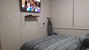 Une chambre avec un lit, un téléviseur fixé au mur et une petite étagère.