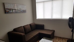 Olohuoneessa on kulmasohva, lasitasoinen sohvapöytä, lattiatuuletin ja seinällä taulu.