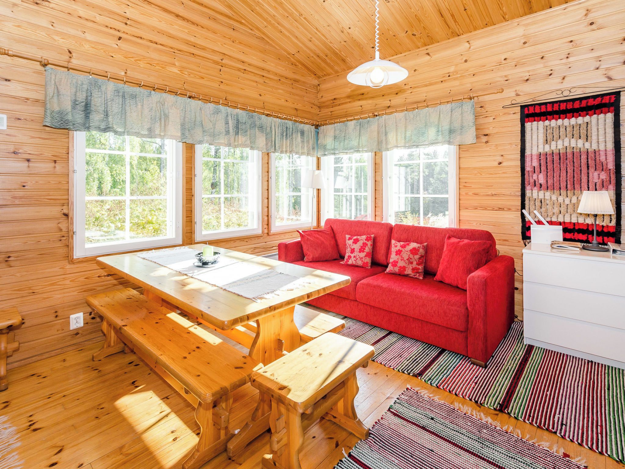 A wooden cabin interior with a red sofa, wooden dining table, benches, and large windows.