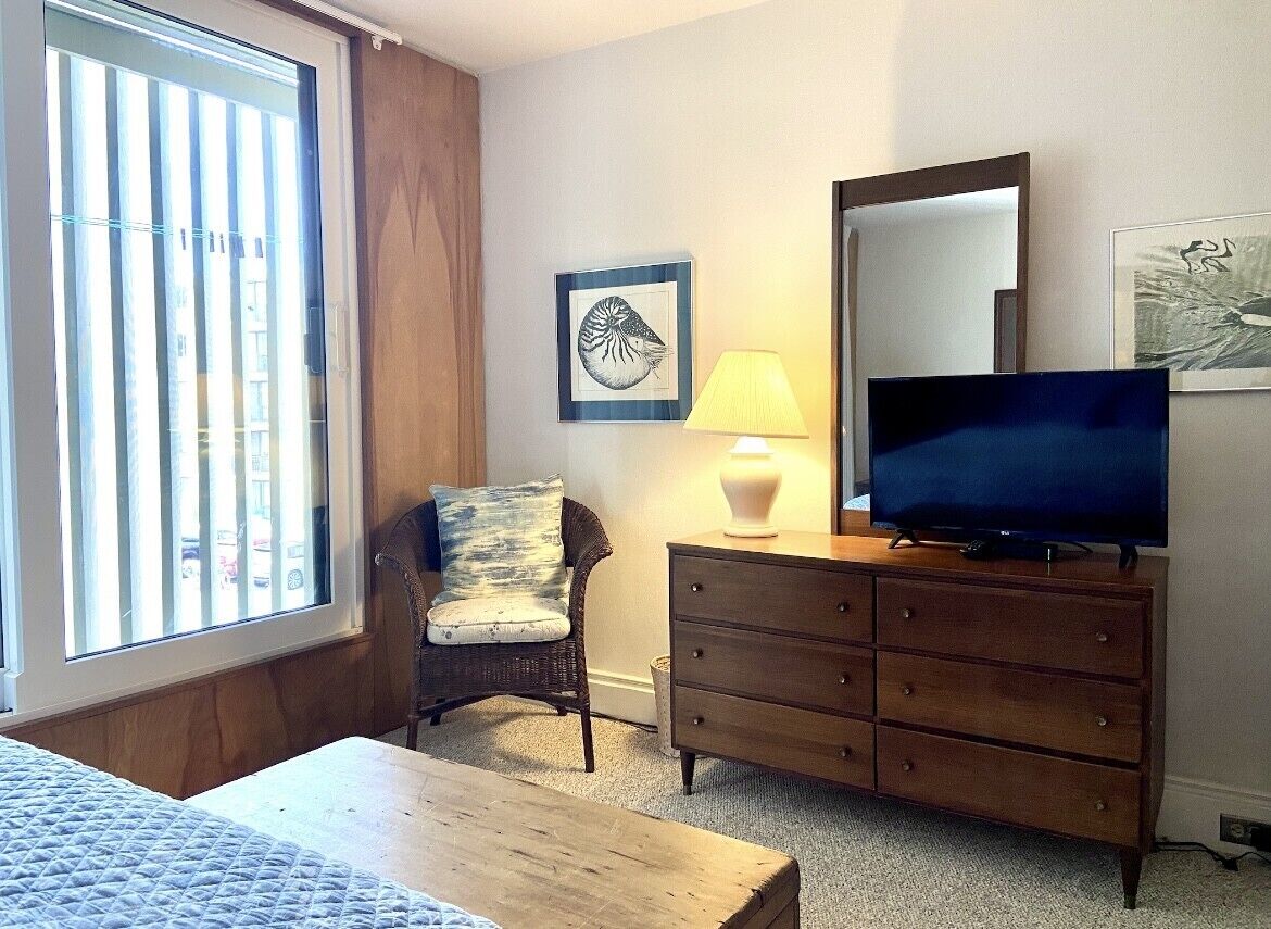 Une chambre d’hôtel comprenant un lit, une commode en bois, un téléviseur, une chaise, une lampe et une fenêtre avec des stores.