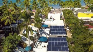 A coastal area with solar panels on buildings, palm trees, and a beach in the background.