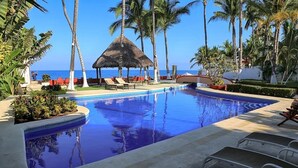 A pool area with lounge chairs, a thatched-roof structure, and palm trees.