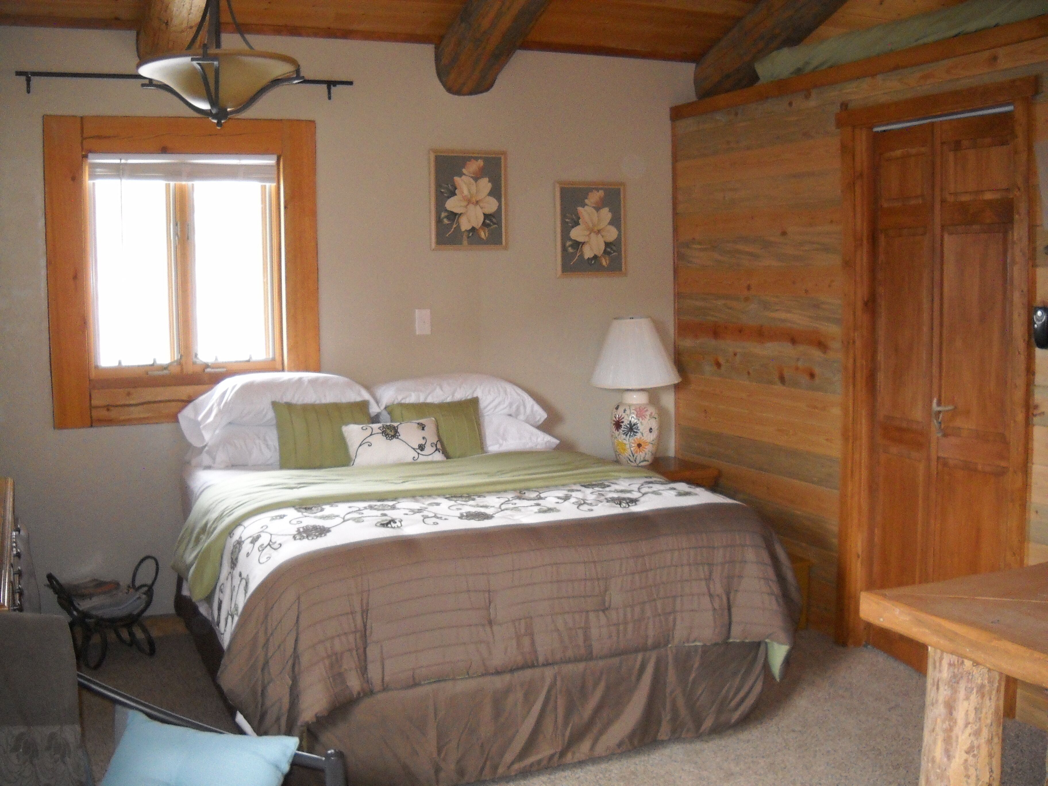 A cozy bedroom with a wooden ceiling, a bed with green and white bedding, a nightstand with a lamp, and two floral wall art pieces.