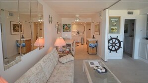 Sala de estar con un sofá, una mesa de centro y una lámpara. Se ve una cocina al fondo con una mesa de comedor y sillas.