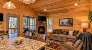 Interior acolhedor de uma cabana com lareira de pedra, móveis de madeira e uma televisão na parede.