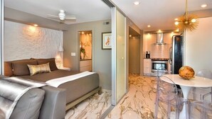 A modern hotel room with a bed, a seating area with a table and chairs, a kitchen area, and a bathroom. - OCEAN FRONT CONDO.  ENJOY YOUR MORNING COFFEE SITTING ON YOUR PRIVATE BALCONY WATCHING THE SUNRISE. (Rehoboth Beach)
