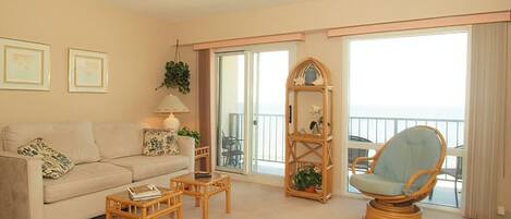 A living room with a sofa, coffee table, and a wicker chair. There is a balcony with a railing and a view of the sea.