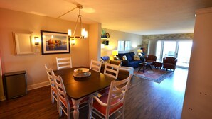 Un comedor con una mesa de madera y sillas blancas, una sala de estar con sofá y sillones, y una cocina con refrigerador.