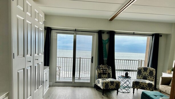 A spacious room with a view of the beach, a balcony with a railing, and two patterned chairs.