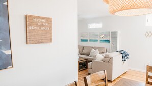 A modern living room with a wooden sign, a sofa, a coffee table, and a dining area with chairs. - 65 Steps to Ocean • Private Home • 2-Car Garage (San Diego)