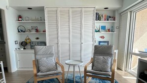 Una habitación con dos sillones, una mesita y un tapete azul estampado. Se ve una estantería con diversos objetos y una ventana con vistas.