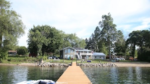 A wooden dock leading to a house by a lake, surrounded by trees and a small boat. - Home of 1000 Island#1-Directly on STL  river, private beach & FREE boat docking  (Gananoque)