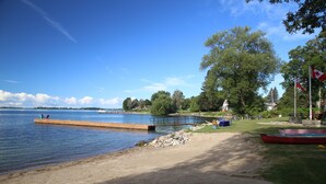A lakeside park with a wooden dock extending into the water, a sandy beach, and a statue in the background. - Home of 1000 Island#1-Directly on STL  river, private beach & FREE boat docking  (Gananoque)
