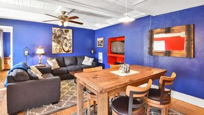 Een woonkamer met een blauwe wand, een plafondventilator, een houten eettafel met stoelen, een zwarte bank met decoratieve kussens, en een bevestigde televisie.