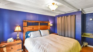Een slaapkamer met een houten hoofdbord, een bed met wit beddengoed, een nachtkastje met een lamp, een wandlamp en een raam met gordijnen.