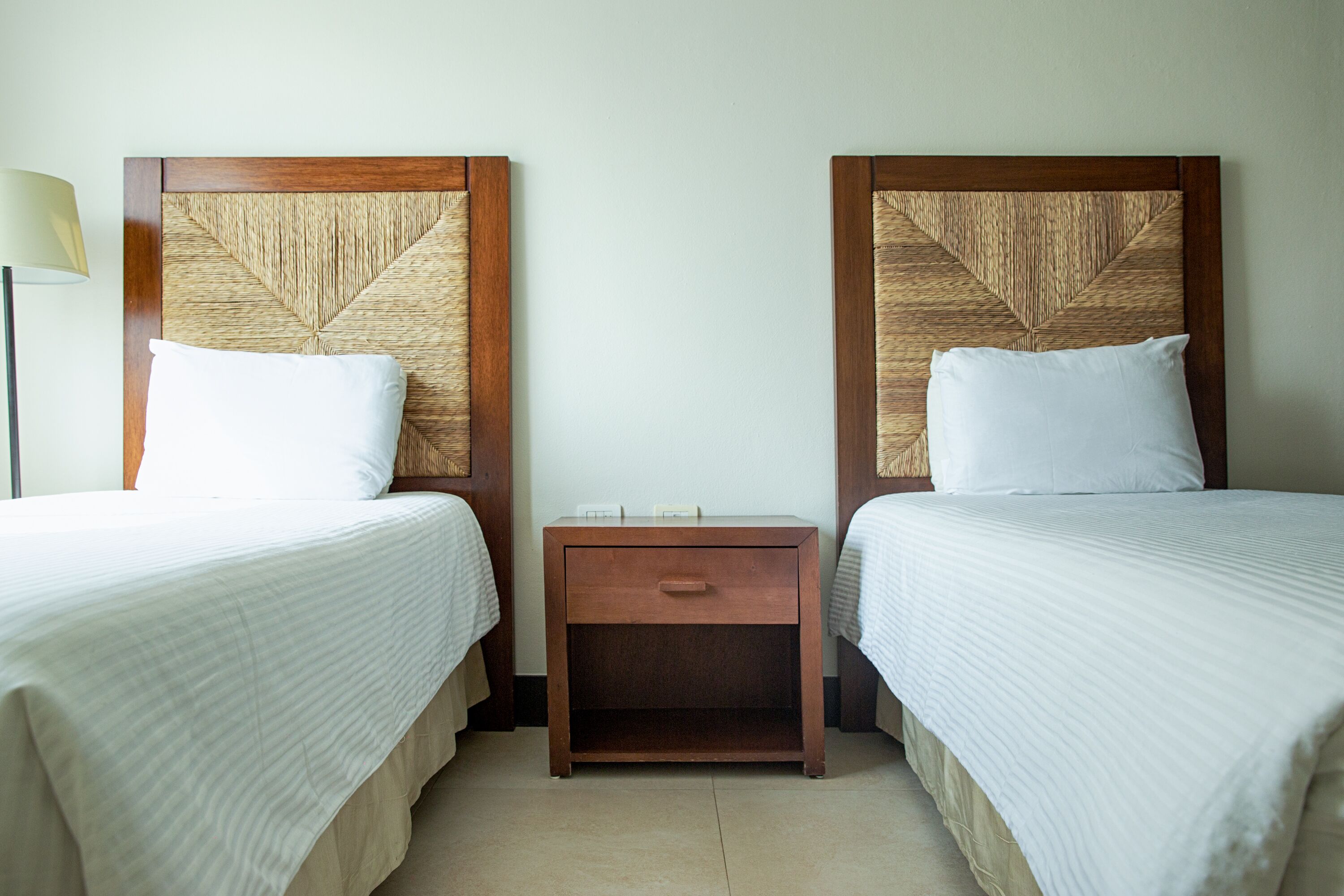 A hotel room with two single beds, a wooden nightstand with a drawer, and a lamp on the left side.