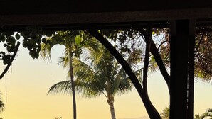 A view from a covered outdoor area with a table and chairs, overlooking a landscaped garden with palm trees and a body of water in the background. - Ekahi 29C, Ocean View,  Steps away from the pool (Wailea)