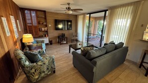 A living room with a sofa, armchair, and a TV. There is a ceiling fan, a window with curtains, and a bookshelf. - Ekahi 29C, Ocean View,  Steps away from the pool (Wailea)
