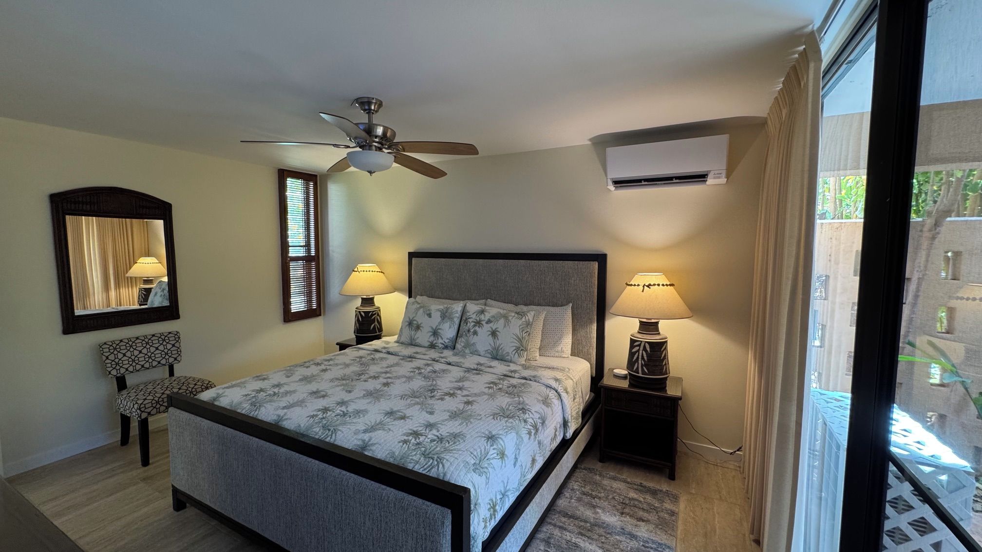 A bedroom with a bed, two bedside tables with lamps, a ceiling fan, a window with blinds, and a wall-mounted air conditioning unit.
