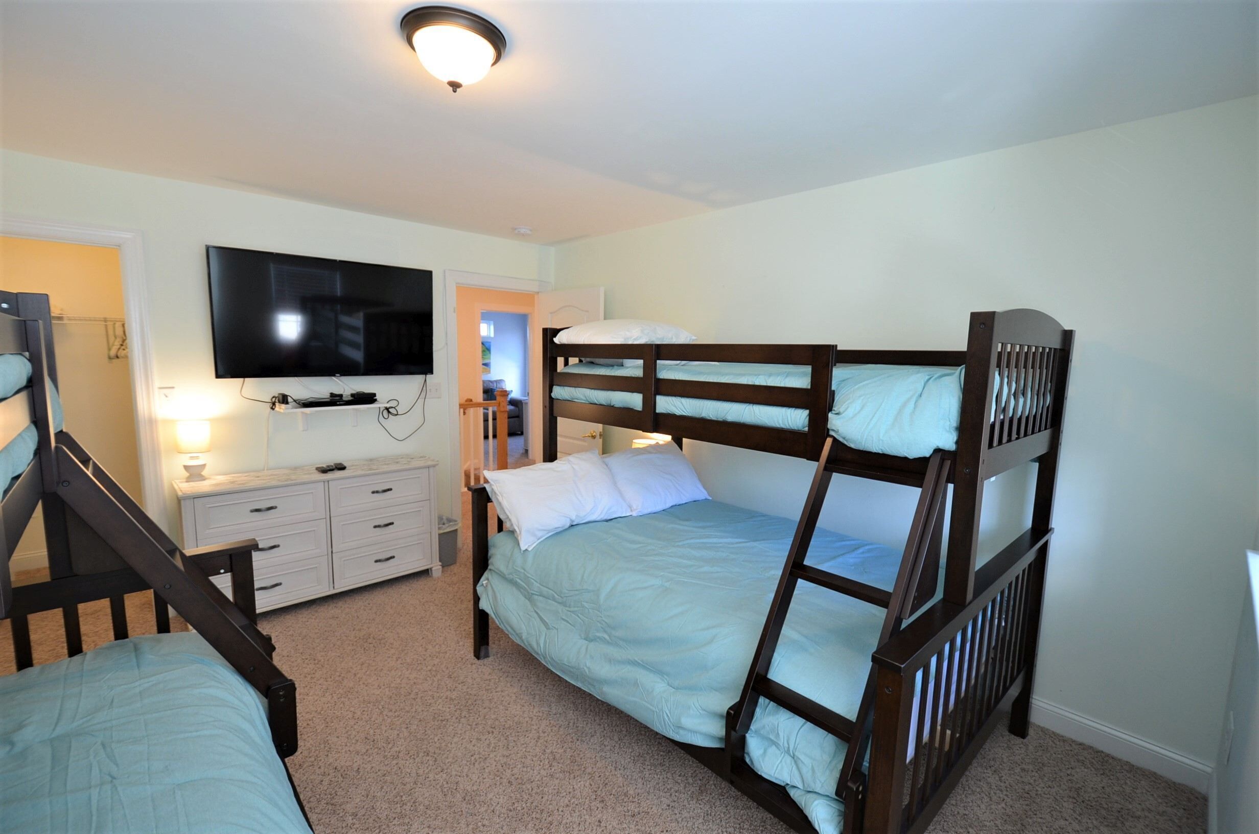 A room with two bunk beds, a flat-screen TV mounted on the wall, a dresser, and a door leading to another room.