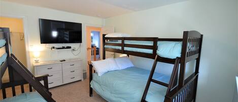 A room with two bunk beds, a flat-screen TV mounted on the wall, a dresser, and a door leading to another room.