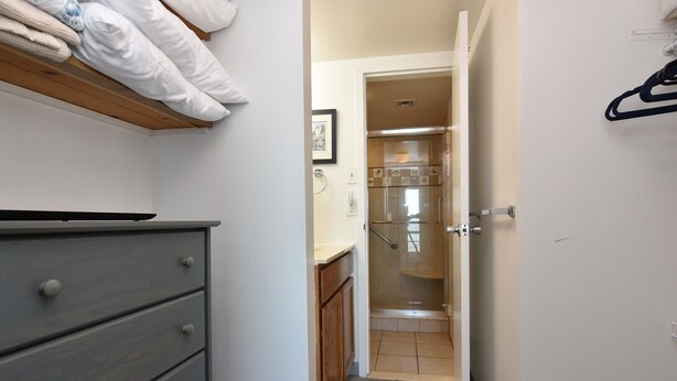 A narrow hallway with a wooden dresser, a bed with white bedding, and a shower area with a glass door.