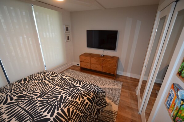 Une chambre à coucher comprenant un lit, un téléviseur fixé au mur, une commode en bois et une fenêtre avec des stores.