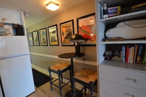 A kitchen with a refrigerator, a coffee maker, a table with a lamp, and a shelf with books and a fish model.