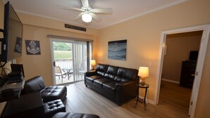 A living room with a leather sofa, a coffee table, a TV, and a ceiling fan. There is a door leading to another room and a sign on the wall. - ENJOY the Community Pool & Tennis Court! (Rehoboth Beach)