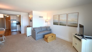 A compact living space with a kitchenette, a grey sofa, a wooden chest, a TV on a cabinet, and three framed pictures on the wall. - Edgewater House - Ocean Front Units With Spectacular Views! (Rehoboth Beach)
