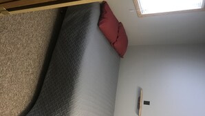 Un lit avec un matelas gris et un oreiller rouge, une tête de lit en bois et un petit objet noir accroché au mur.