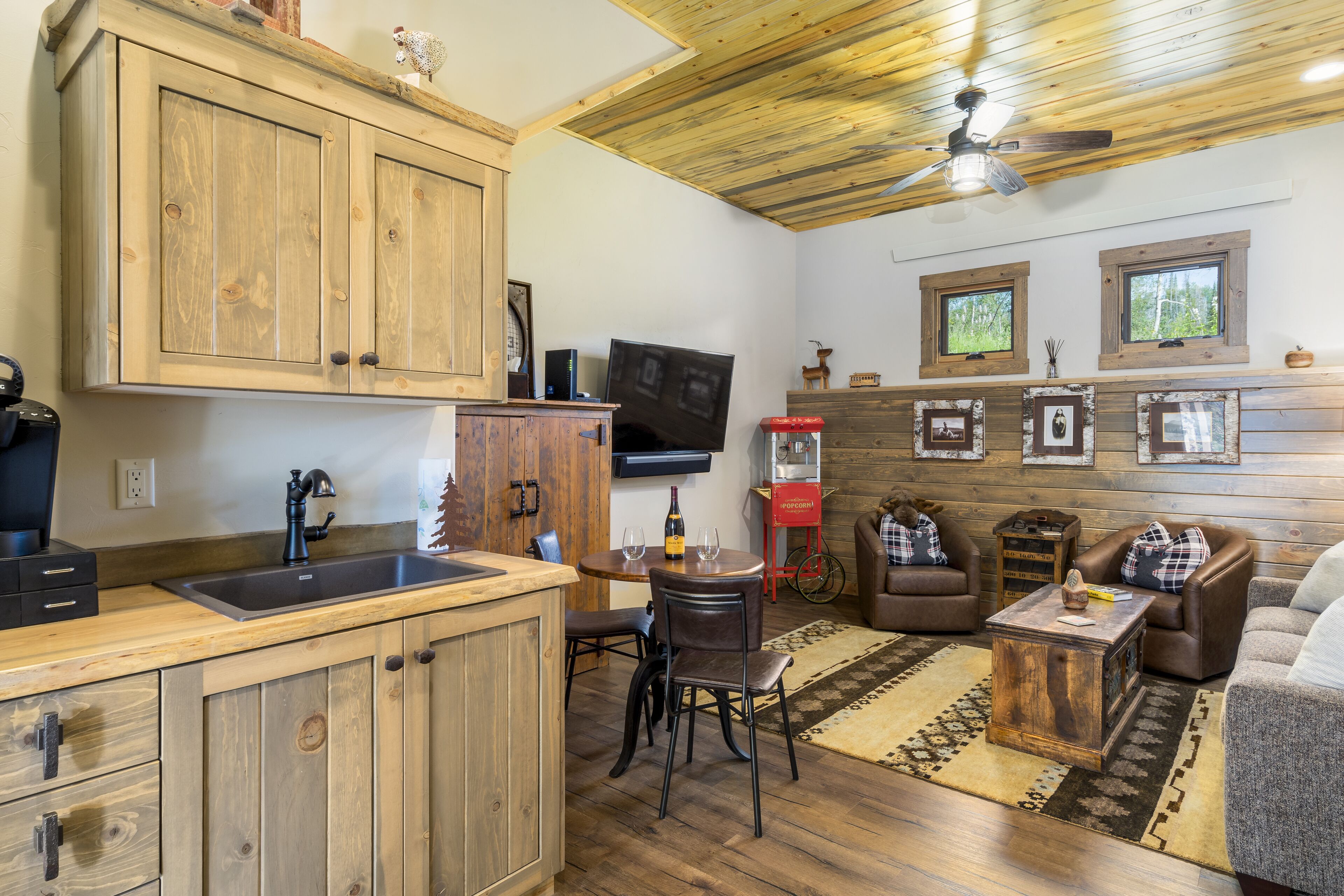 Une cuisine rustique avec des meubles en bois, un évier et un coin repas comprenant une table et des chaises. On y trouve également un ventilateur de plafond, un téléviseur et une fenêtre donnant sur les arbres.