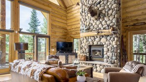 Interior de una cabaña de troncos con chimenea de piedra, muebles de cuero y un televisor.