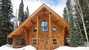 Una cabaña de troncos con un ventanal grande, un porche y el suelo cubierto de nieve.