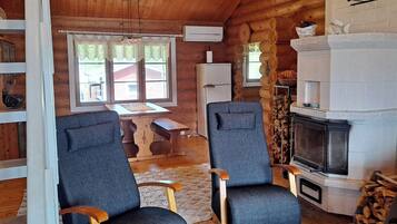 A cozy cabin interior with two armchairs, a wooden dining table, a fireplace, and a window with curtains.