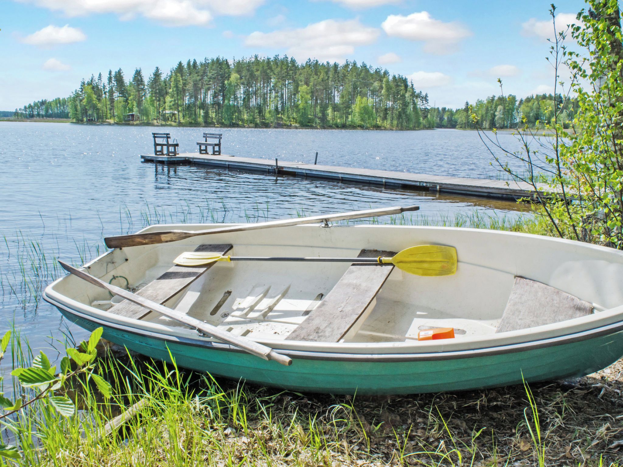 Un bote con remos en un lago tranquilo, amarrado cerca de una orilla cubierta de hierba y un bosque al fondo.