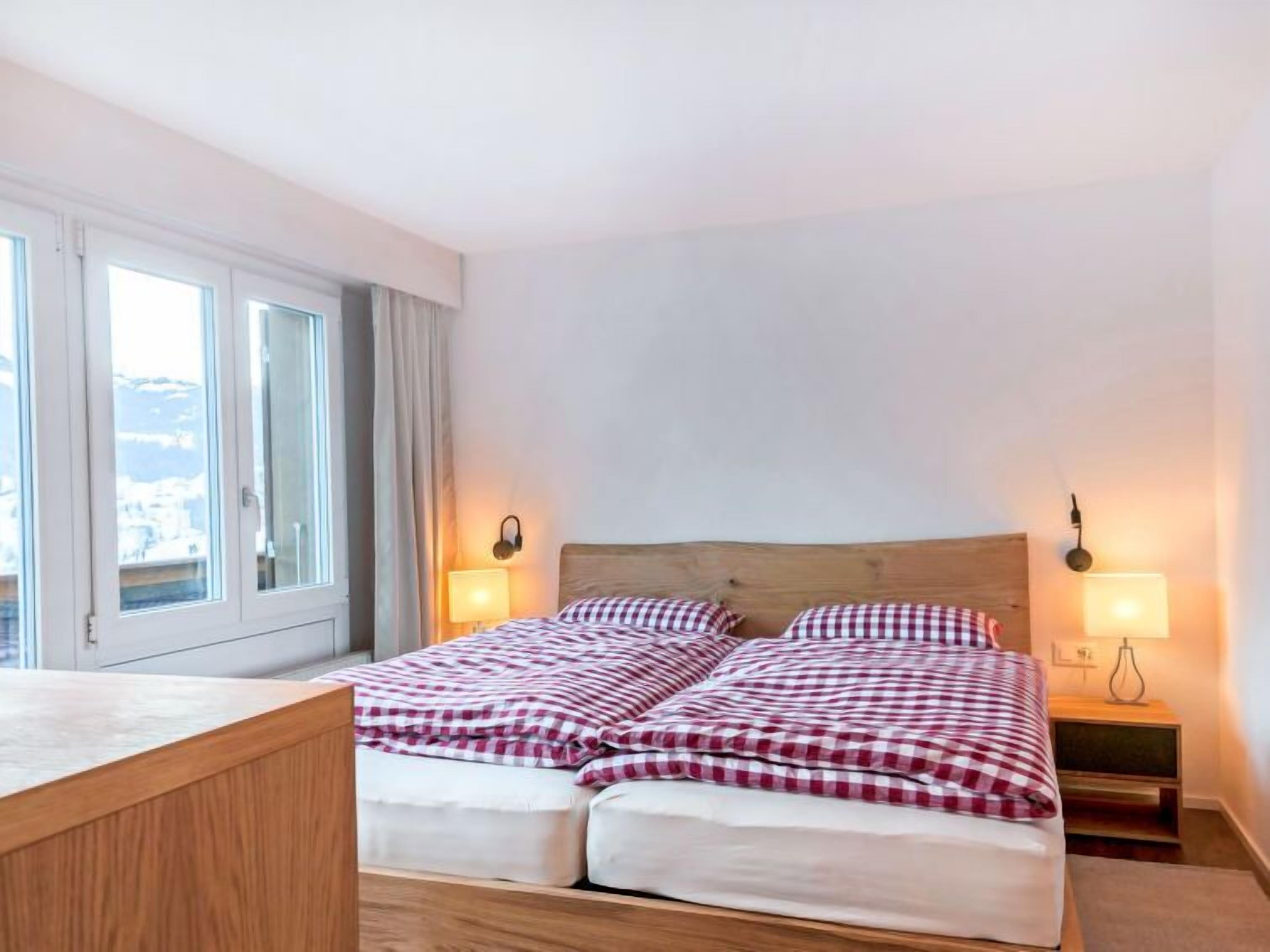 Una cama doble con colcha a cuadros, dos lámparas de noche y un ventanal con cortinas.