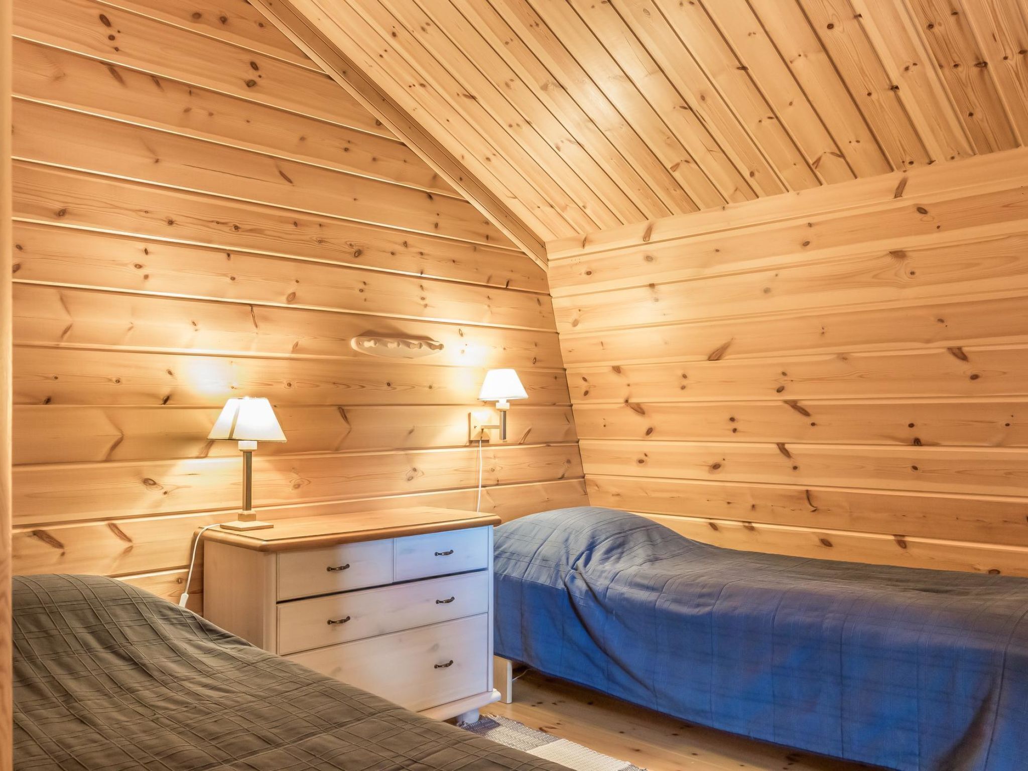 A cozy cabin room with wooden walls, a bed, a nightstand with drawers, and two wall-mounted lamps.
