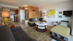 A modern living room with a sofa, a round coffee table, a bed, a TV, and a kitchen in the background. - Edgewater House - Ocean Front Units With Spectacular Views! (Rehoboth Beach)