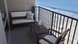 Ein Balkon mit einer Holzbank, einem kleinen Tisch und einem Glasbehälter, mit Blick auf den Strand.
