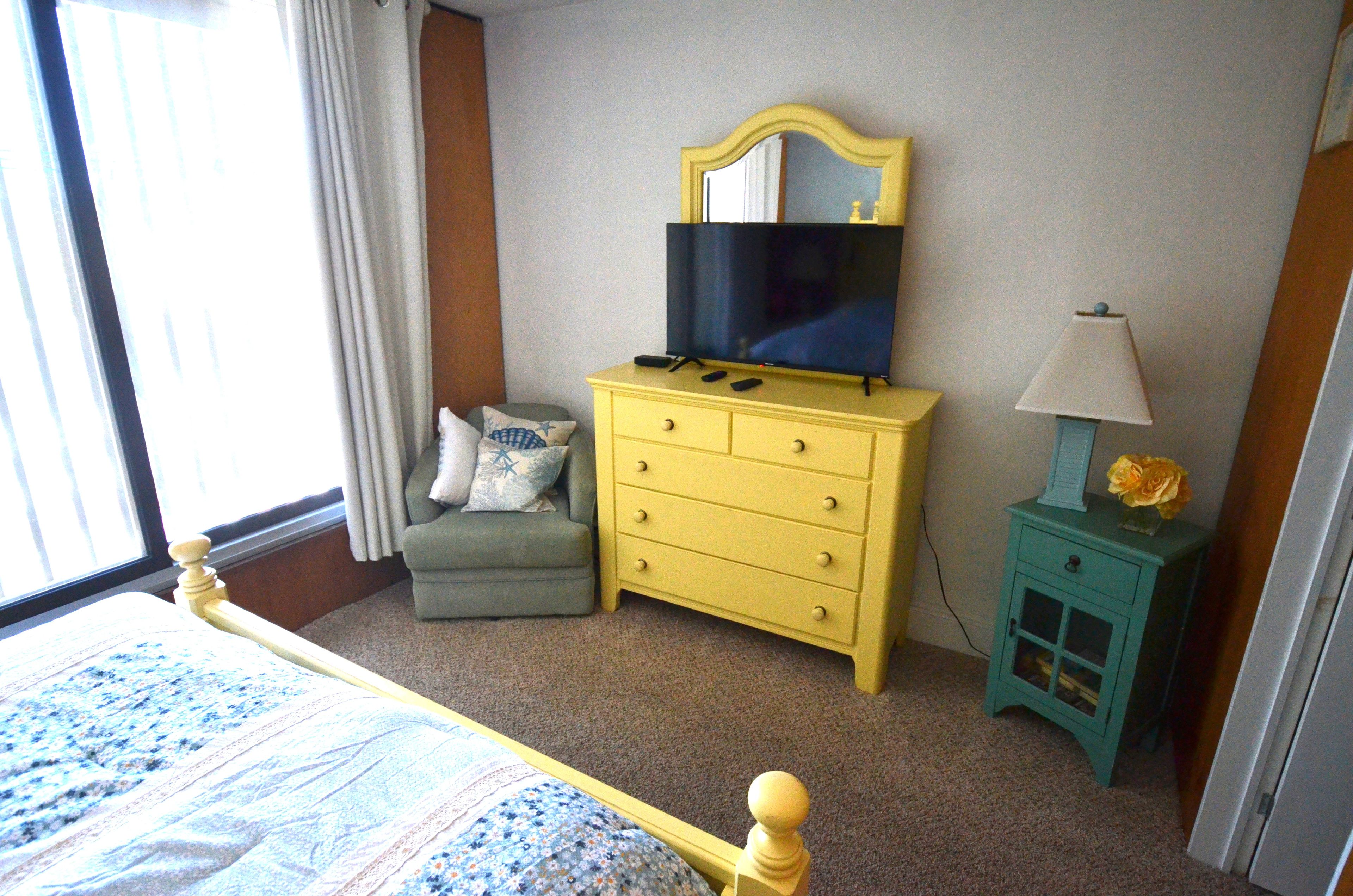 A hotel room with a bed, a yellow dresser, a television, a small table with a lamp, and a window with curtains.